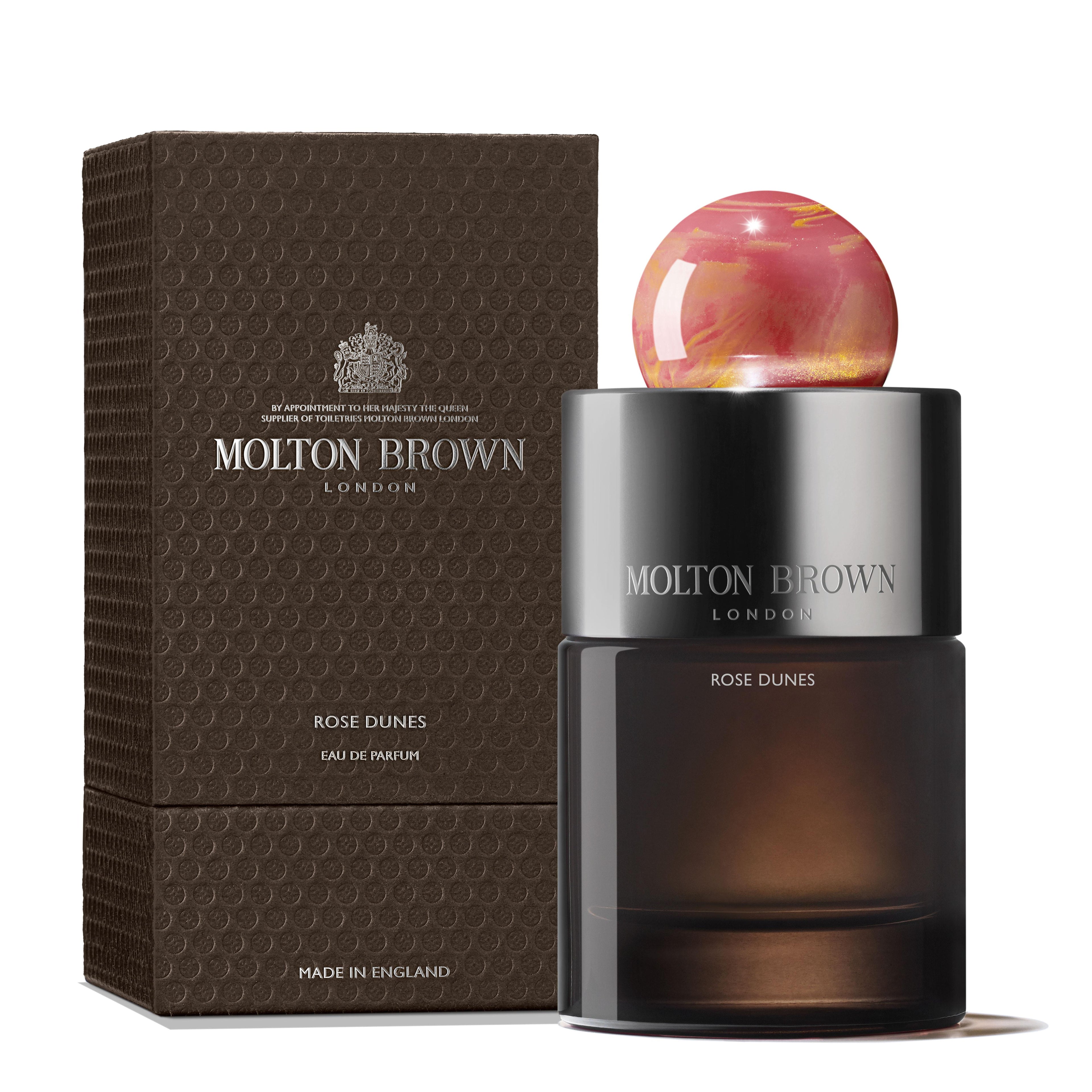 A bottle of MoltonBrown Cyprus's Rose Dunes Eau de Parfum 100ml is displayed alongside its textured brown packaging. The bottle is distinguished by a round, marbled pink-and-gold cap and a dark base, showcasing the MoltonBrown logo and Rose Dunes name. This fragrance captures a luxurious oudh accord with notes of spicy saffron.