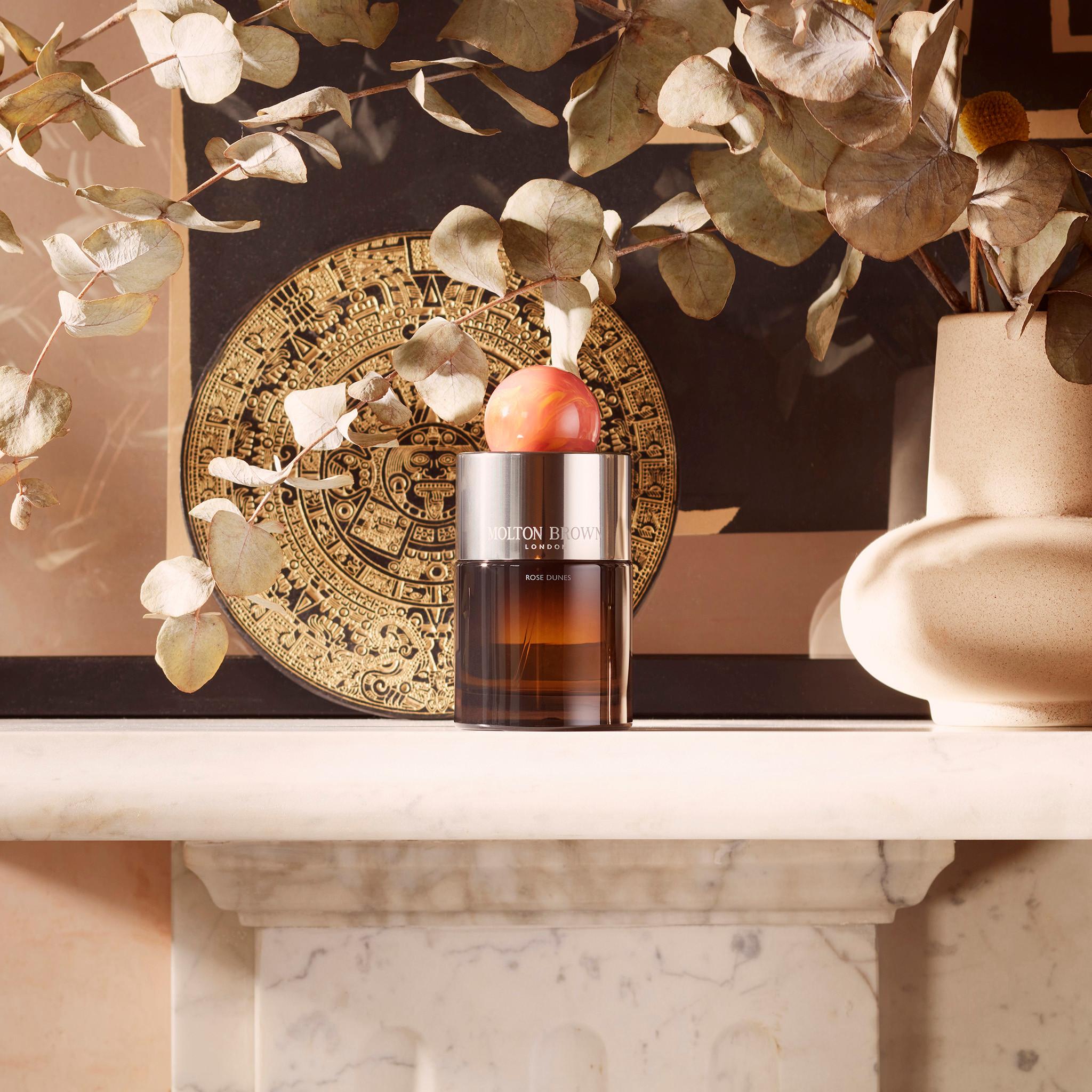 A decorative setup featuring a brown glass bottle of MoltonBrown Cyprus' Rose Dunes Eau de Parfum (100ml) with a label on a marble surface. Surrounding it are spicy saffron-scented eucalyptus branches, a beige vase, and a circular artistic plate with intricate designs. Soft, natural lighting enhances the warm tones.