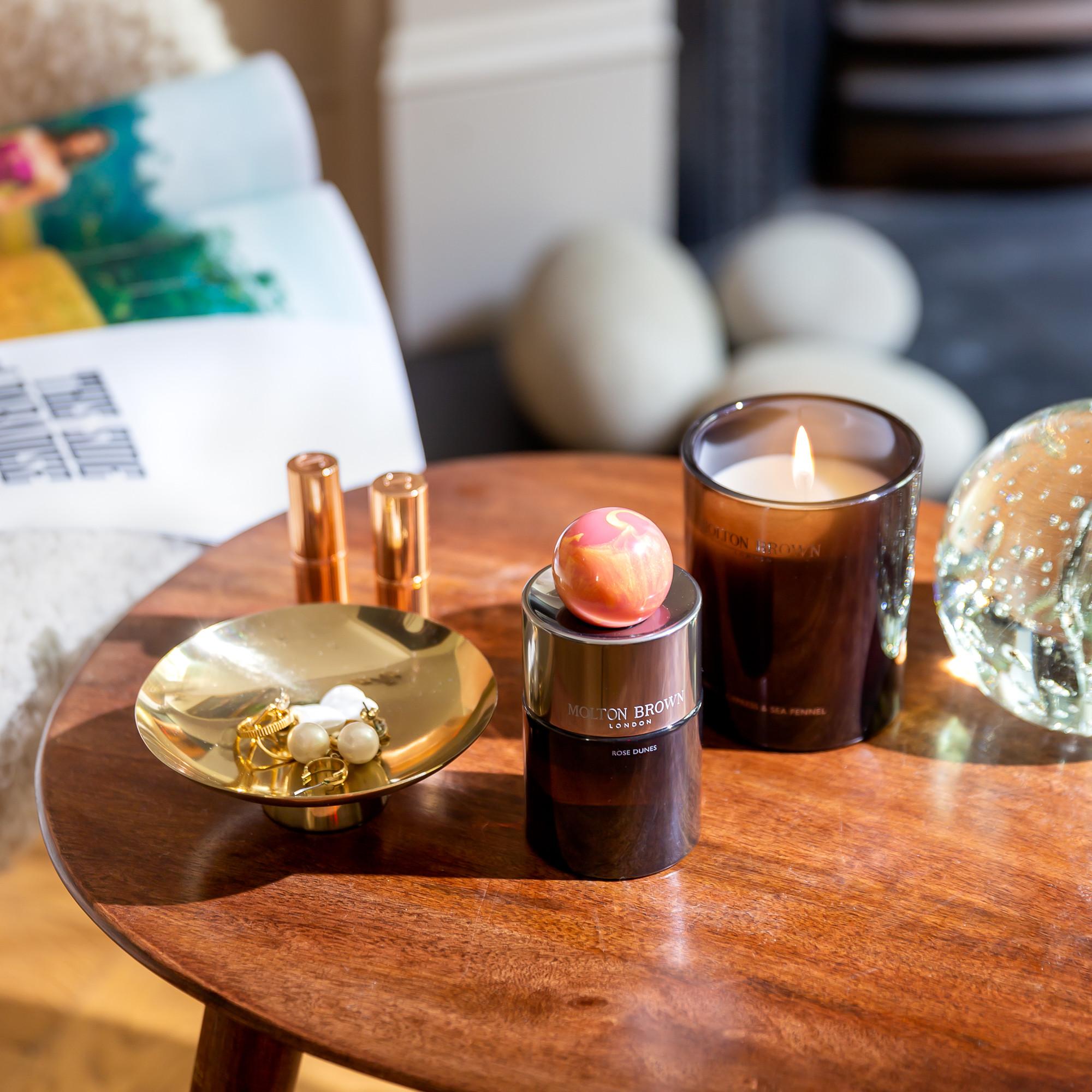 A wooden table holds an opulent oudh accord scented candle, a jar with apple-shaped soap, a gold dish with jewelry, two copper lipstick tubes, and a decorative glass orb. A cozy seat with a colorful pillow and MoltonBrown Cyprus's Rose Dunes Eau de Parfum 100ml is partially visible in the background.