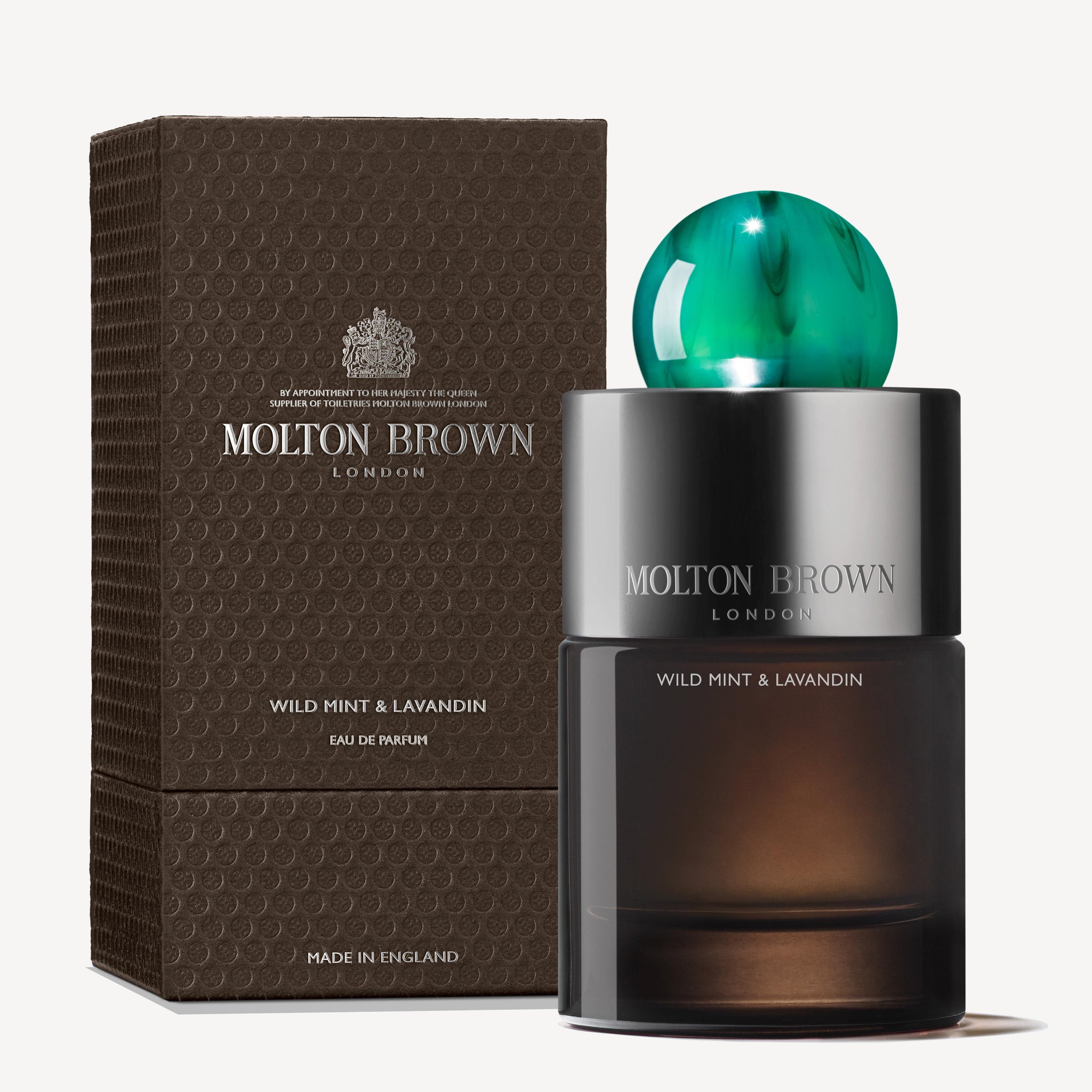 The Wild Mint & Lavandin Eau de Parfum from MoltonBrown Cyprus features a chic bottle adorned with a green spherical cap. It comes alongside an elegant textured brown box highlighted with silver accents and branding, introducing a subtle touch of lavender to its sophisticated presentation.