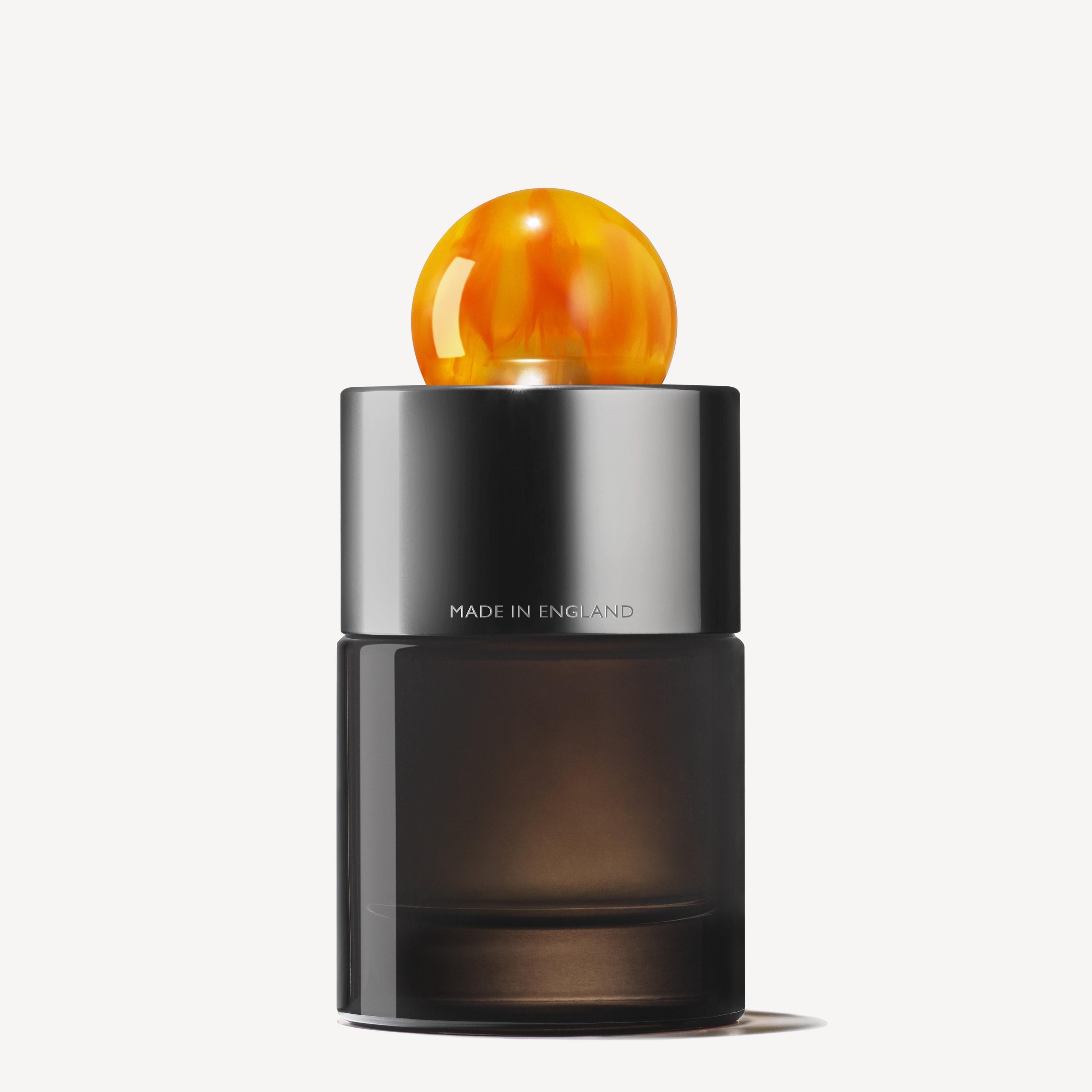 The Sunlit Clementine & Vetiver Eau de Parfum 100ml by MoltonBrown Cyprus features a sleek design with a dark, cylindrical base and a shiny metallic middle section inscribed with "Made in England." It's crowned with a bright, round orange cap that resembles amber. This citrus fragrance combines notes of clementine and mandarin against a plain white background.