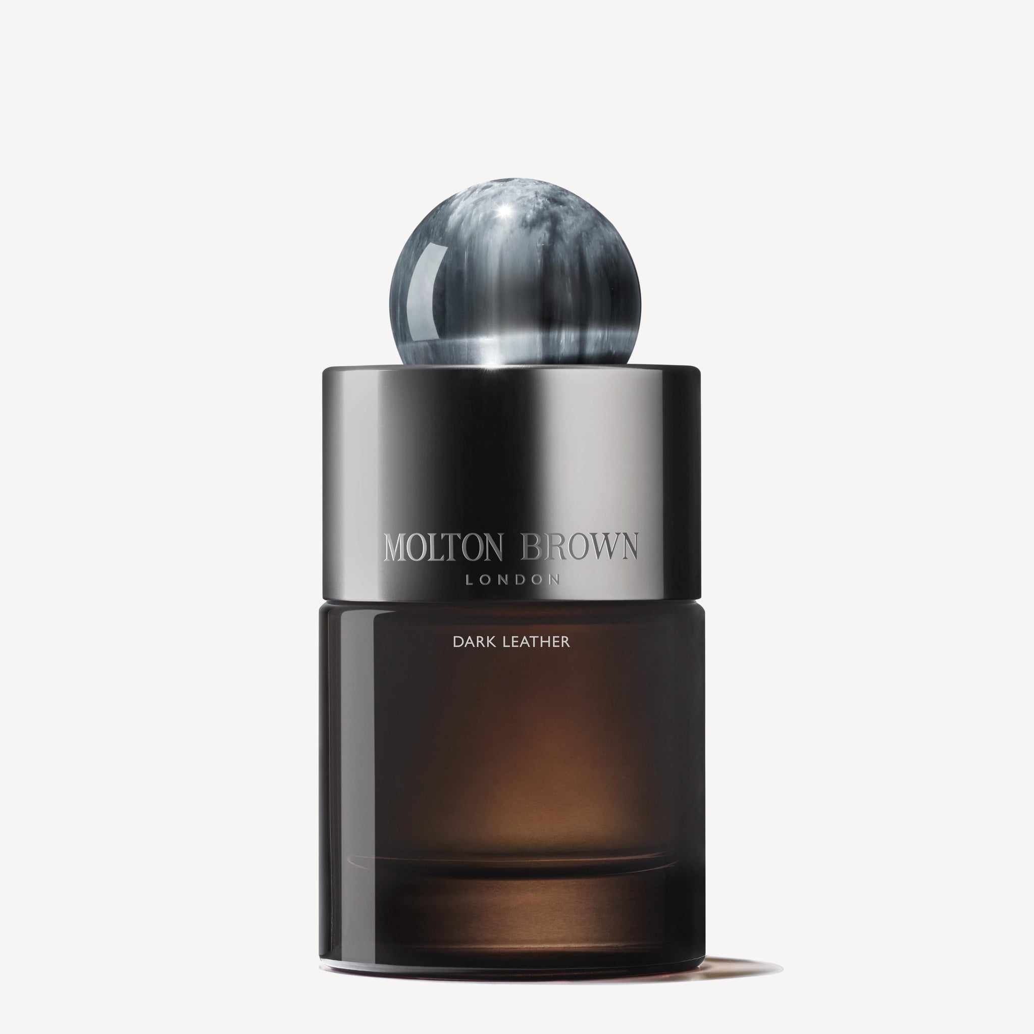 The Dark Leather Eau de Parfum 100ml from MoltonBrown Cyprus features a sleek, dark design with a reflective sphere cap and a gradient glass base. It is infused with smoky leather notes, while the brand name is elegantly printed in white on a silver band at the top.