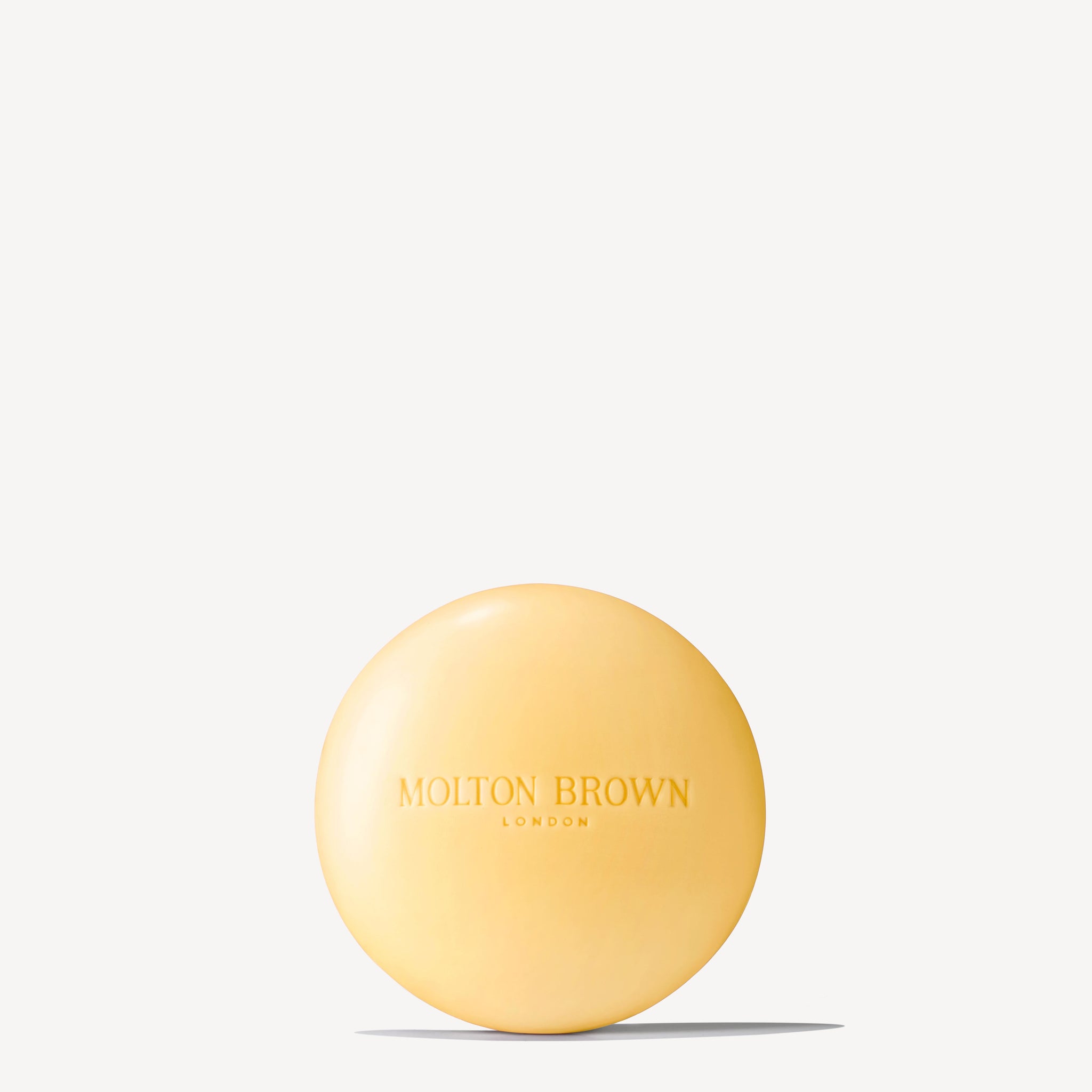 A round, yellow Orange & Bergamot Perfumed Soap from MoltonBrown Cyprus, adorned with the brand logo on the front. Featuring a refreshing citrus fragrance, it is positioned against a plain gray background and casts a subtle shadow beneath.