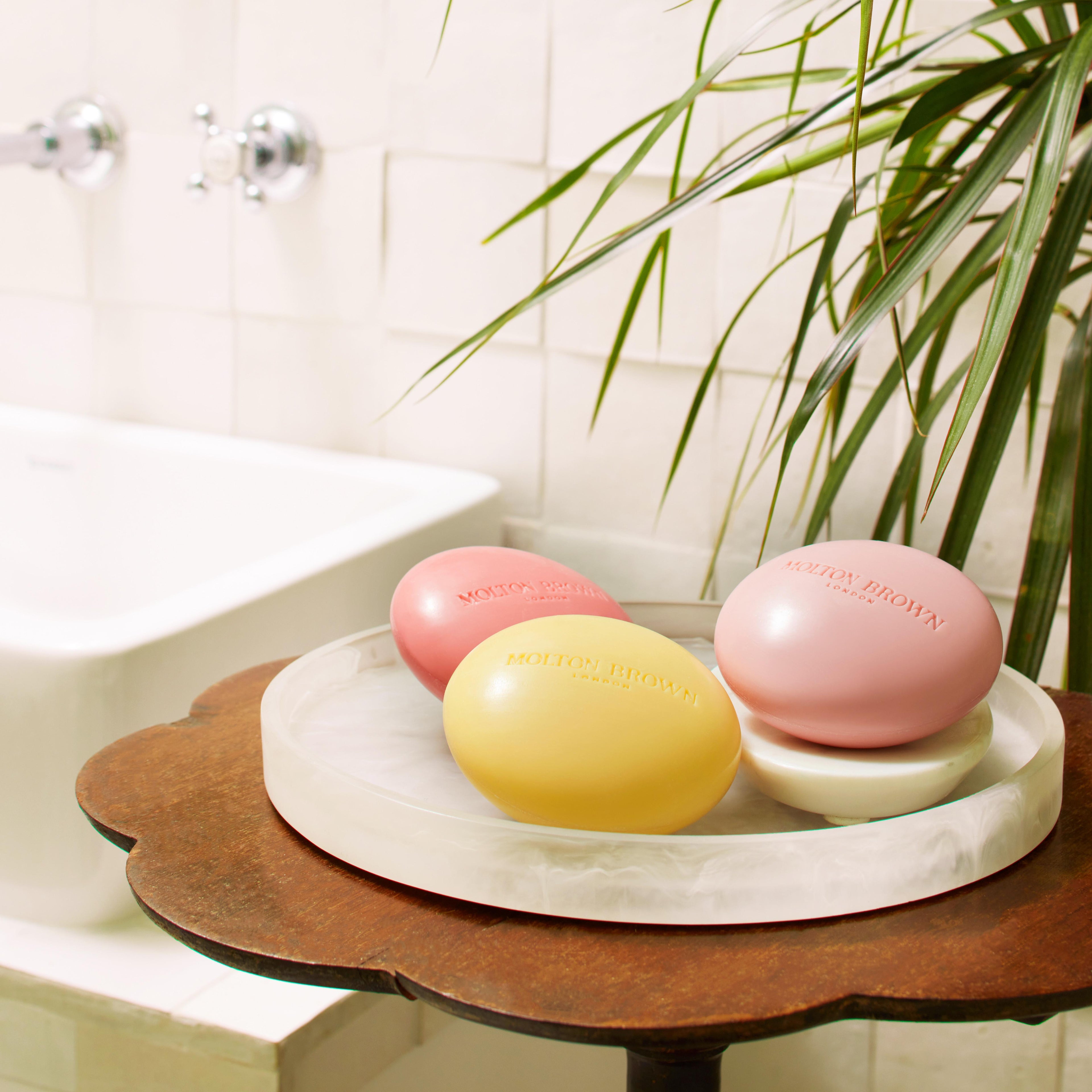 A bathroom setting showcases a round, marble platter on a wooden table, displaying three oval bars of Orange & Bergamot Perfumed Soap by MoltonBrown Cyprus. The soaps, infused with shea butter, come in pastel shades of pink, yellow, and rose. A green plant is nearby, while a white bathtub invites relaxation in the background.