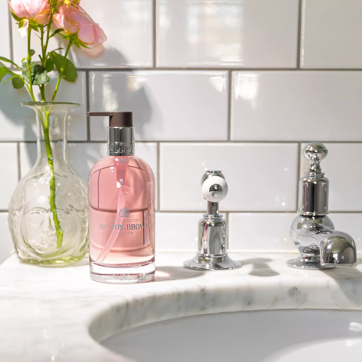 The MoltonBrown Cyprus Delicious Rhubarb & Rose Fine Liquid Hand Wash in a recyclable glass bottle adorns a white marble sink with chrome fixtures, while a vase with pink roses adds charm against the white subway tile wall.