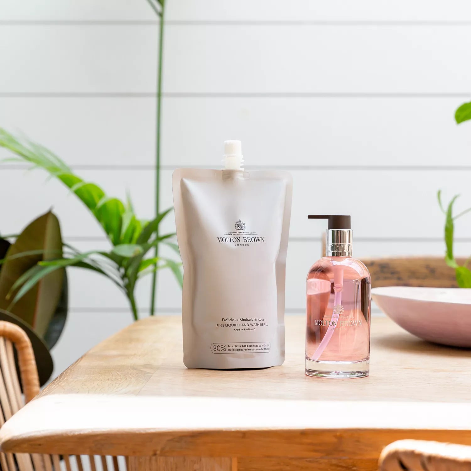 A MoltonBrown Cyprus Delicious Rhubarb & Rose Fine Liquid Hand Wash 200ml glass bottle and refill pouch, both recyclable, rest on a wooden table. Bright space with green plants, light wall backdrop; a pink bowl and glass bottle peek from the right, enhancing the scenes charm.