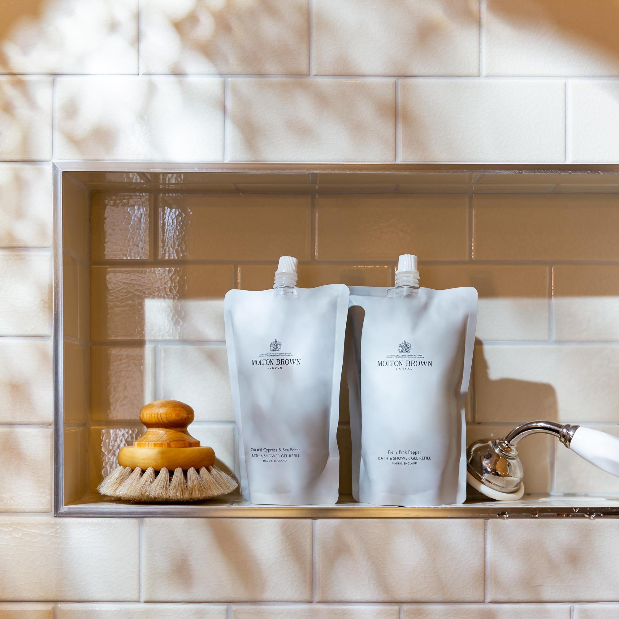 A pair of refill pouches labeled MoltonBrown Cyprus, featuring the Fiery Pink Pepper Bath & Shower Gel Refill 400ml, are placed on a bathroom shelf. They're joined by a bristle brush with a wooden handle and a silver showerhead, as decorative shadows play across the white tiled wall.