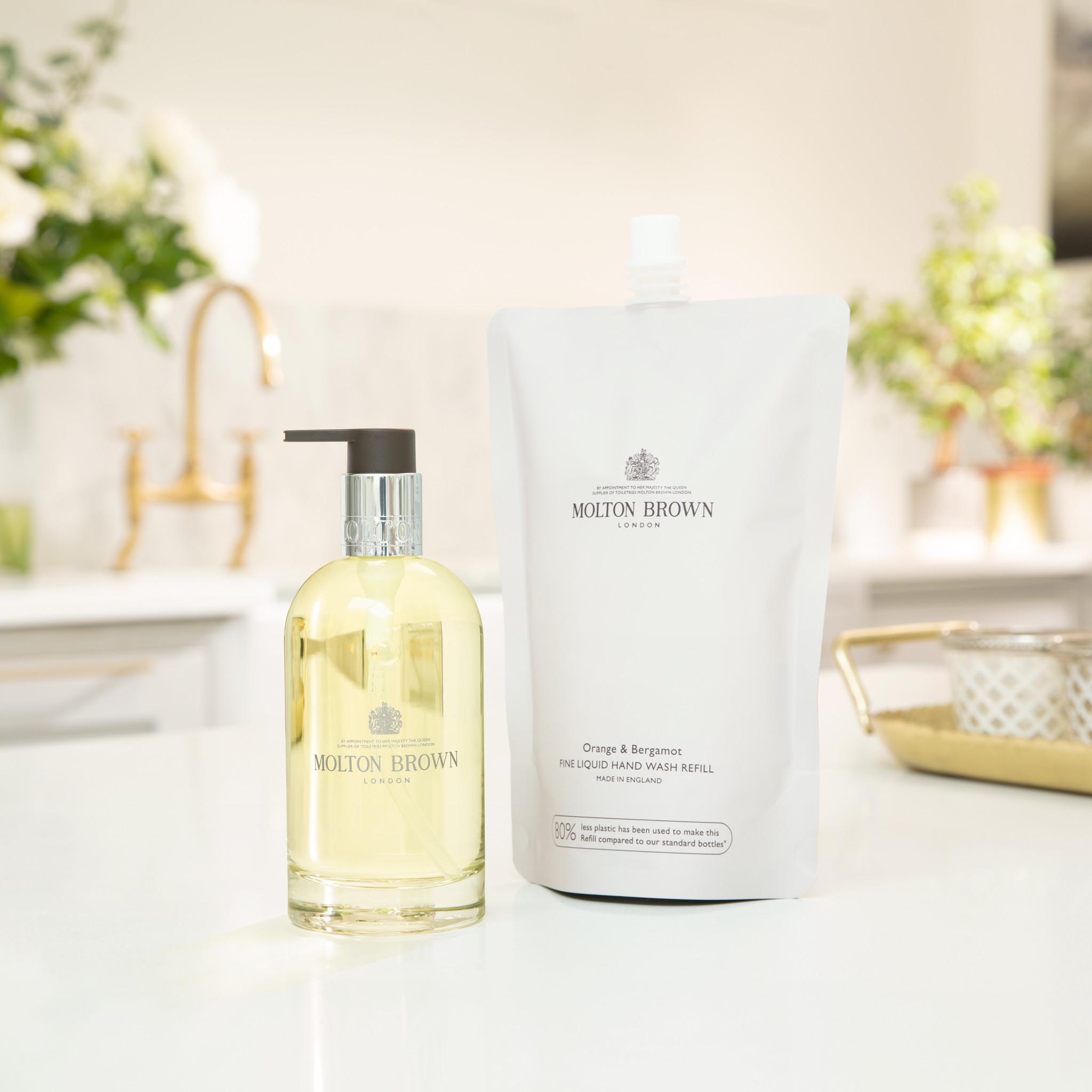 A bottle of MoltonBrown Cyprus Orange & Bergamot Fine Liquid Hand Wash sits alongside its 400ml refill pouch on a kitchen counter, showcasing the vibrant yellow liquid with its signature scent. In the blurred background, flowers accentuate a gold faucet, emphasizing your dedication to sustainable elegance in the kitchen.