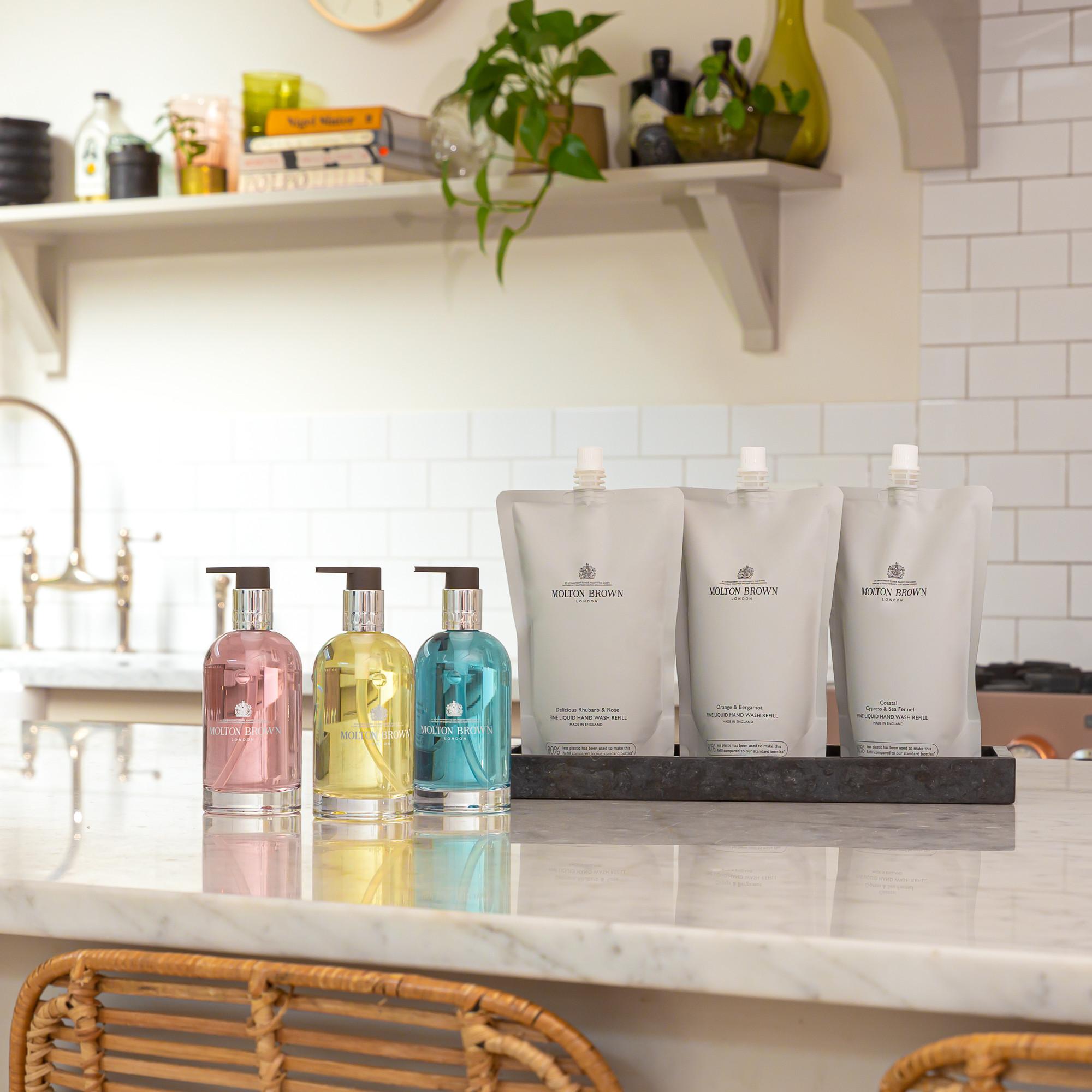 Bottles and refill pouches of MoltonBrown Cyprus's sustainable Orange & Bergamot Fine Liquid Hand Wash Refill 400ml grace the kitchen counter. They sit harmoniously with wicker chairs against a backdrop of white subway tiles, accented by a shelf adorned with decorative items and lush plants.