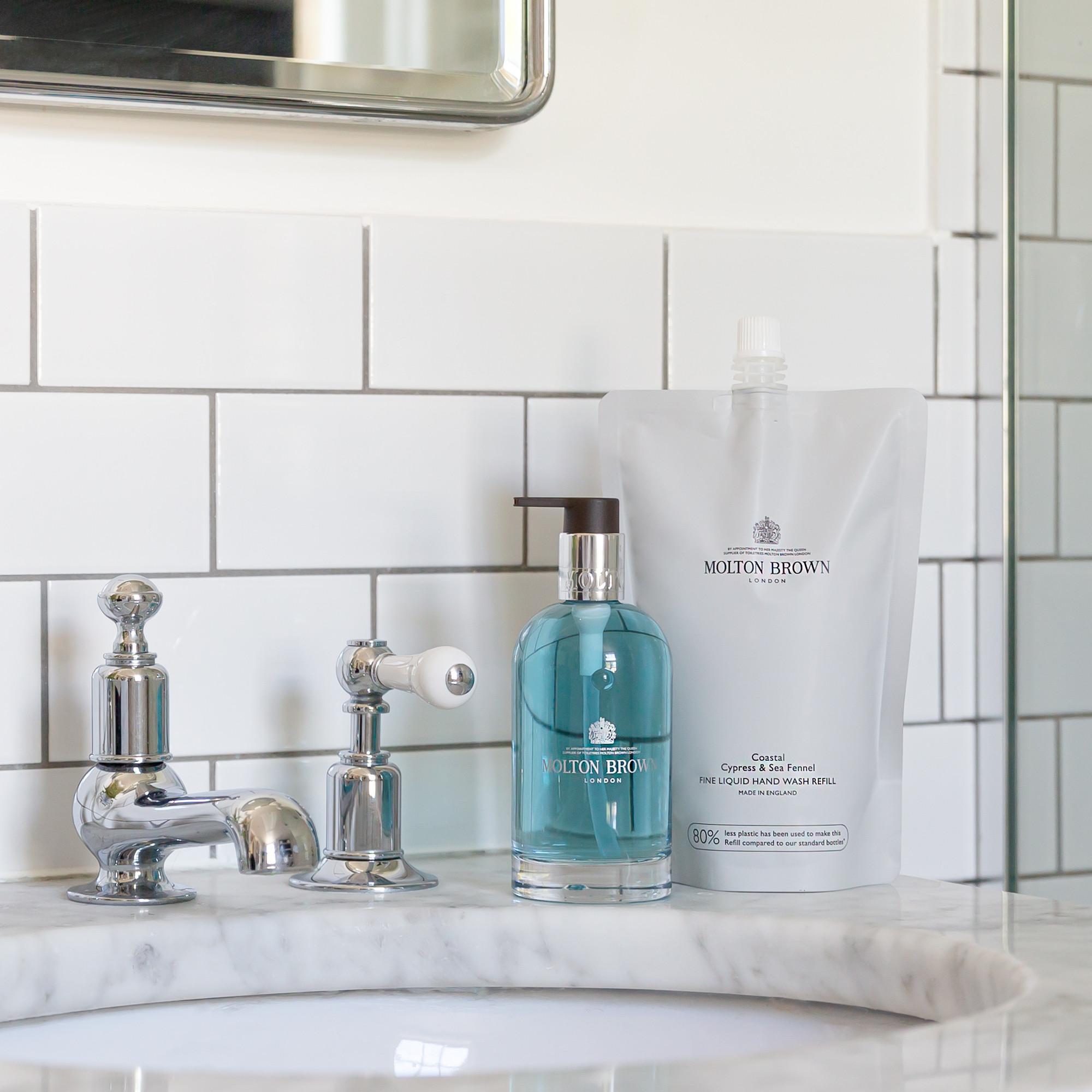 On a marble bathroom sink with a chrome faucet, set against white subway tiles, sits a bottle and refill pouch of MoltonBrown Cyprus Coastal Cypress & Sea Fennel Fine Liquid Hand Wash 400ml, blending luxury with sustainable choices through its marine-inspired fragrance.