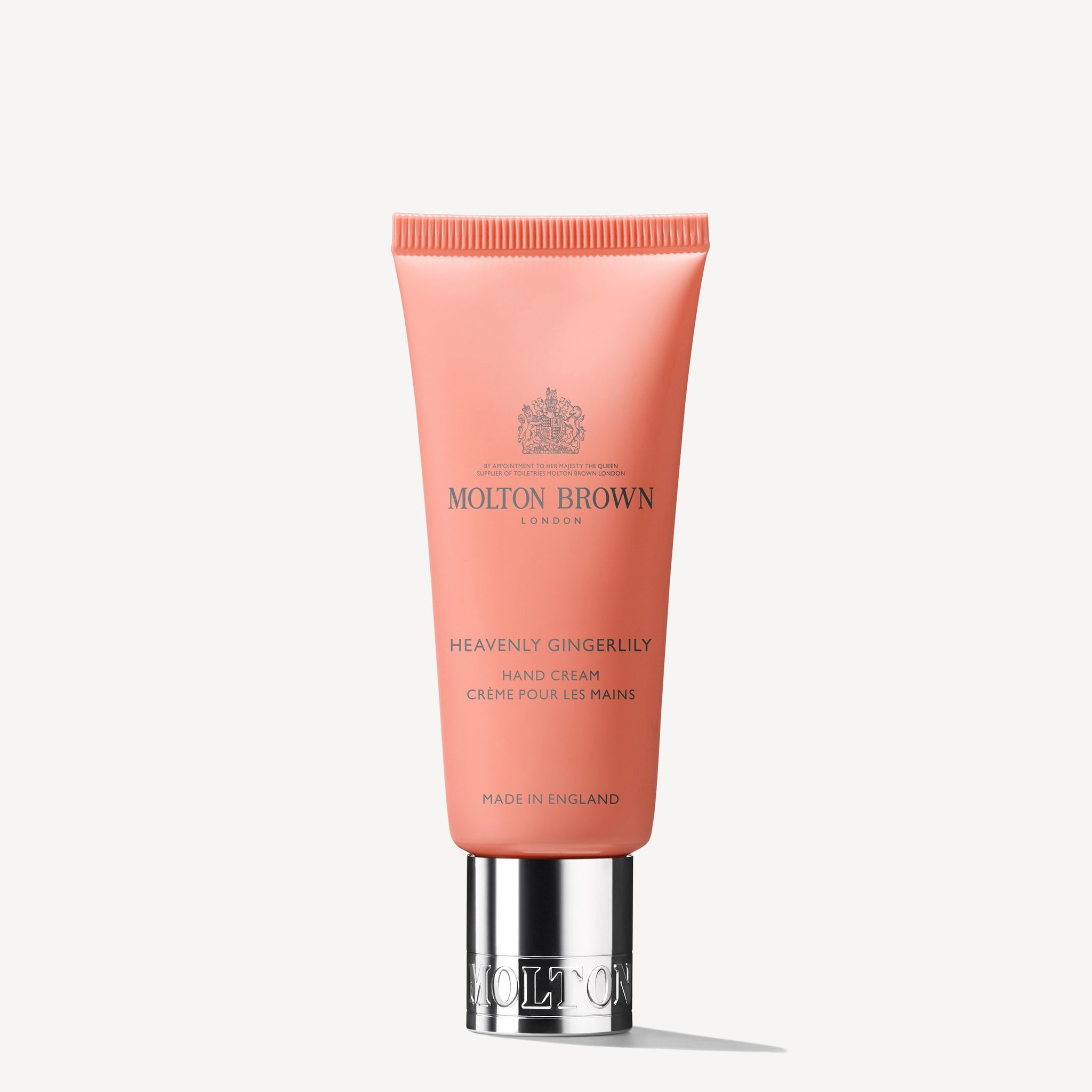 A tube of MoltonBrown Cyprus Heavenly Gingerlily Hand Cream in a pink-orange shade with a silver cap, stands upright against a plain white background. The fragranced tube, made in England, promises moisture and elegance.