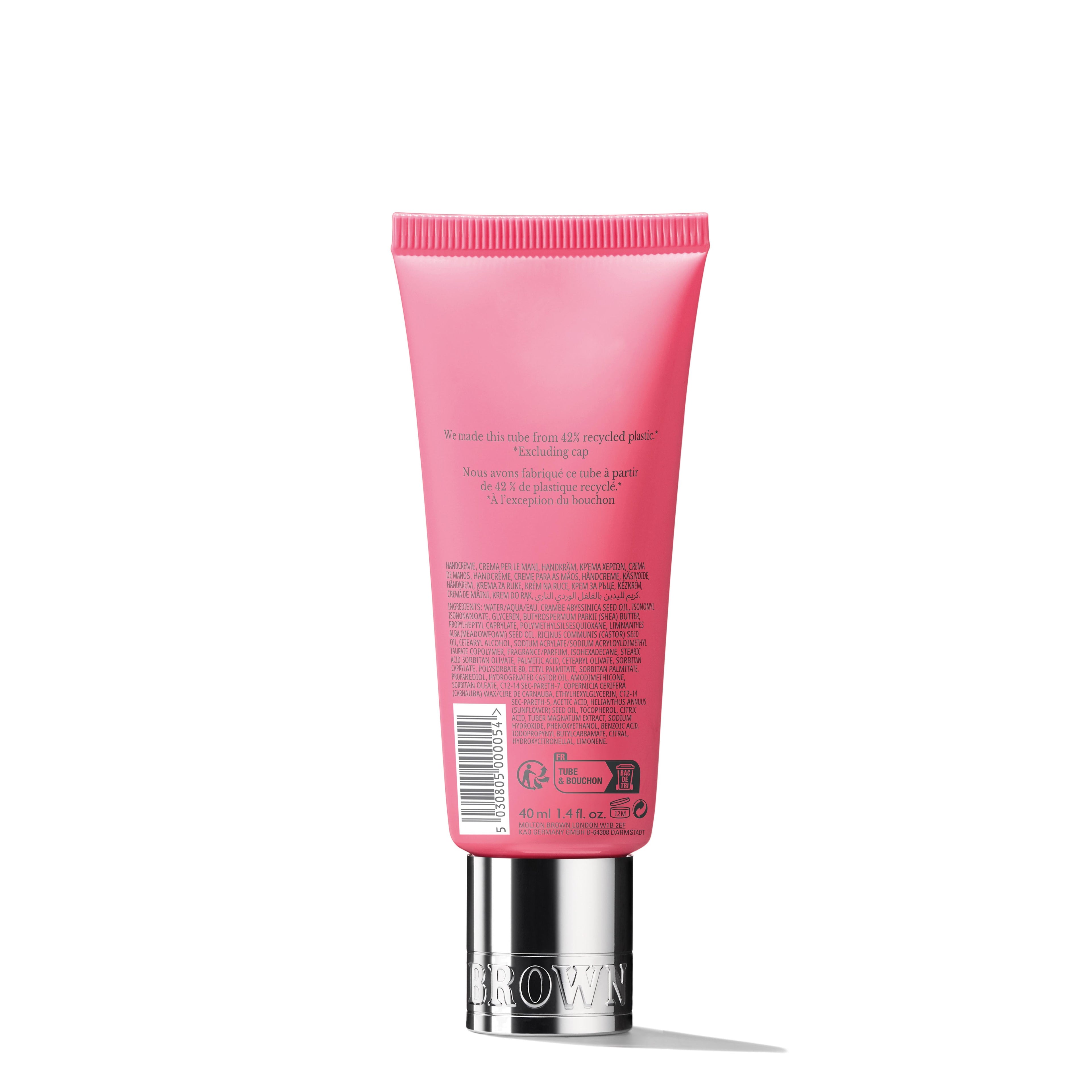 A tube of Fiery Pink Pepper Hand Cream from MoltonBrown Cyprus stands upright, showcasing its pink, opaque packaging with a silver cap. The back displays text and symbols in various languages that provide product information and recycling details. The cream is subtly infused with hints of ginger and pink pepper. The background is plain white.