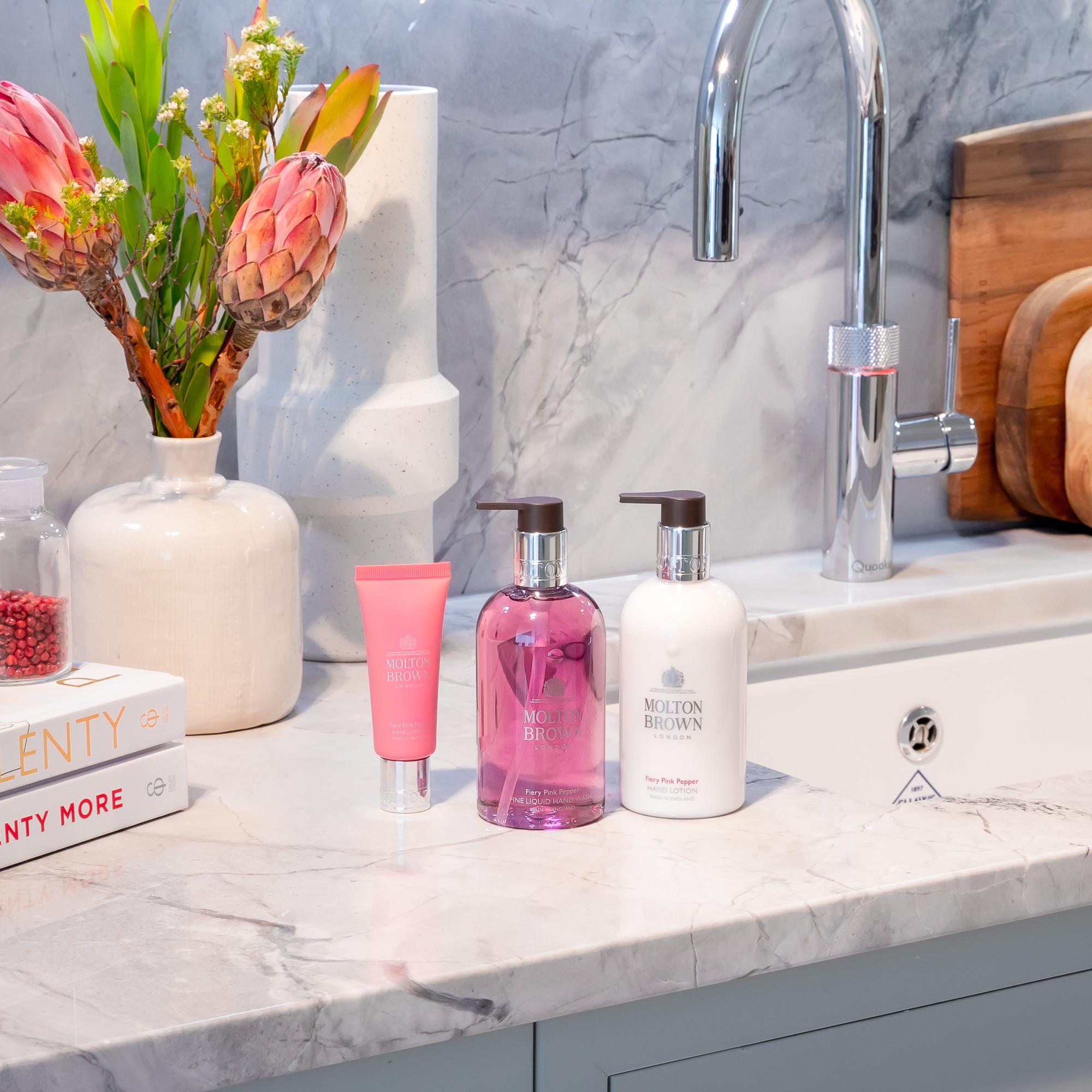 A marble bathroom countertop featuring a silver faucet. On the counter, there are bottles of Molton Brown soap and lotion from MoltonBrown Cyprus, a Fiery Pink Pepper Hand Cream 40ml in a pink tube, a vase filled with protea flowers, cutting boards, and a candle displaying partial "TWENTY MORE" text.