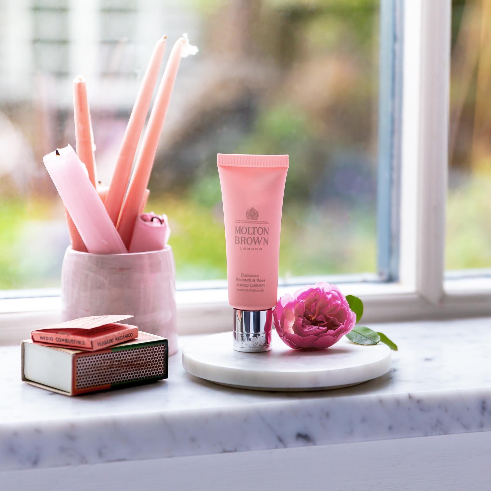 A tube of MoltonBrown Cyprus Delicious Rhubarb & Rose Hand Cream 40ml is elegantly placed on a marble surface by the window, accompanied by a pink rose and matches. Nearby, a cup contains pink candles, with the lush rhubarb leaf garden gently out of focus in the background.