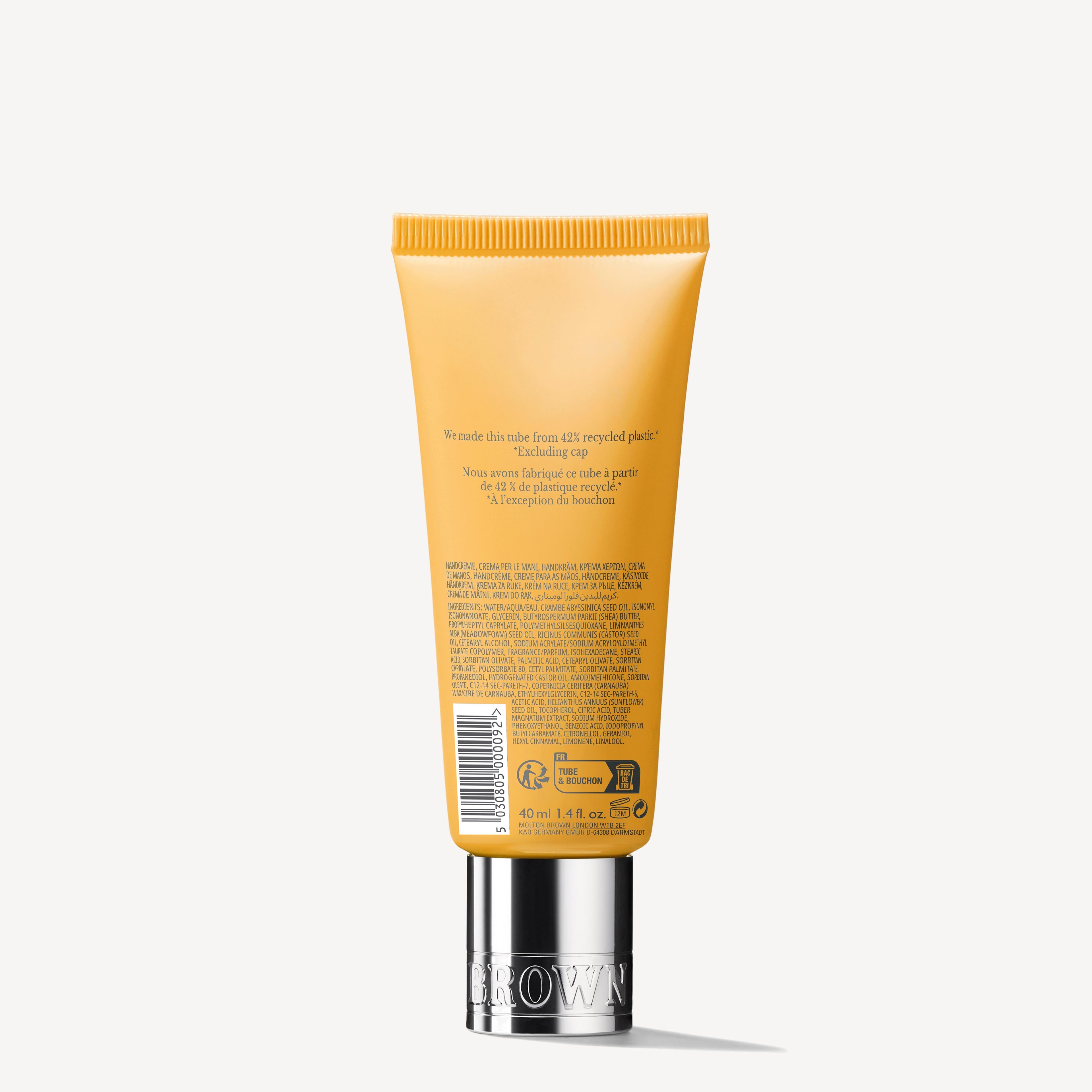 A Molton Brown Cyprus Flora Luminare Hand Cream in an orange tube with a silver cap stands upright, featuring hydration benefits. Text on the back provides product information in multiple languages, and the tube highlights its sustainable packaging made from recycled plastic.