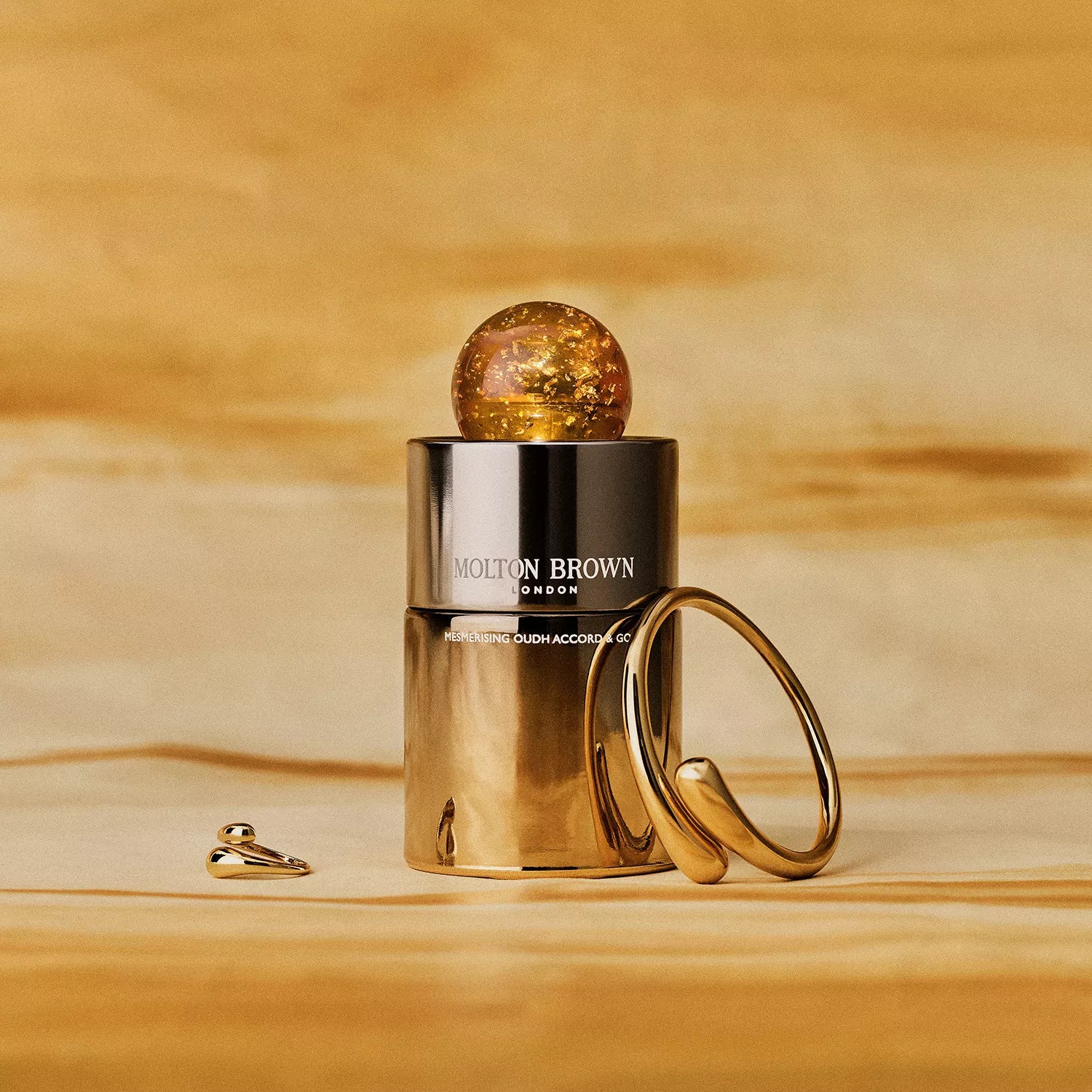 A Limited Edition Mesmerising Oudh Accord & Gold Eau de Parfum 100ml by MoltonBrown Cyprus, featuring a gold metallic bottle and round amber cap, is shown beside two gold jewelry pieces on a gold-toned background.