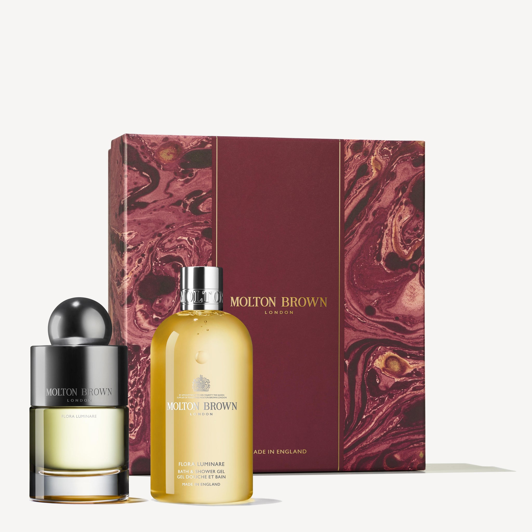 A dark brown Molton Brown gift box sits beside two bottles: a yellow Flora Luminare bath and shower gel with a silver cap and an Eau de Toilette round perfume bottle with a black and silver cap, both featuring MoltonBrown Cyprus branding, from the Flora Luminare Fragrance Gift Set.