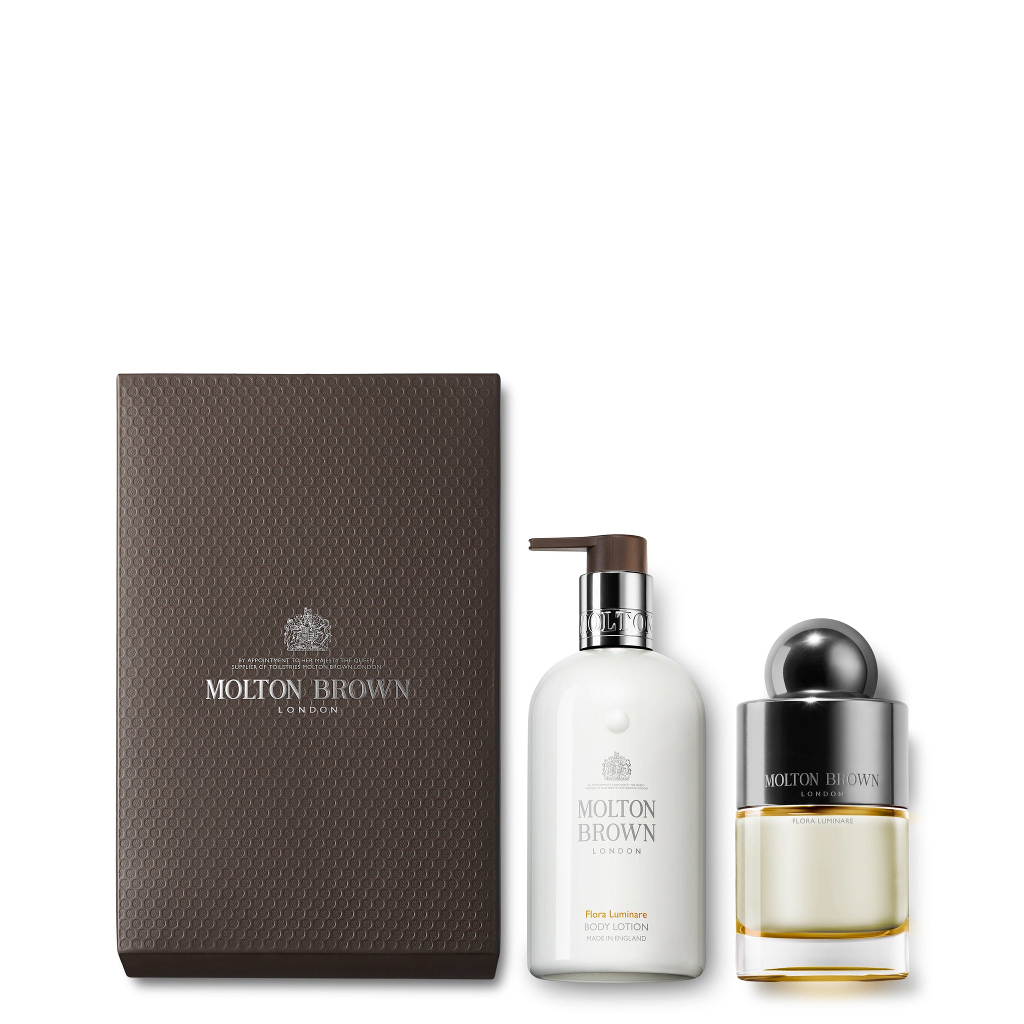 The 100ml Flora Luminare Fragrance Lotion Set from MoltonBrown Cyprus includes a textured brown box on the left, a centrally placed white bottle of Fiery Pink Pepper body lotion equipped with a pump, and a round glass bottle of Polynesian frangipani perfume featuring a silver cap on the right.
