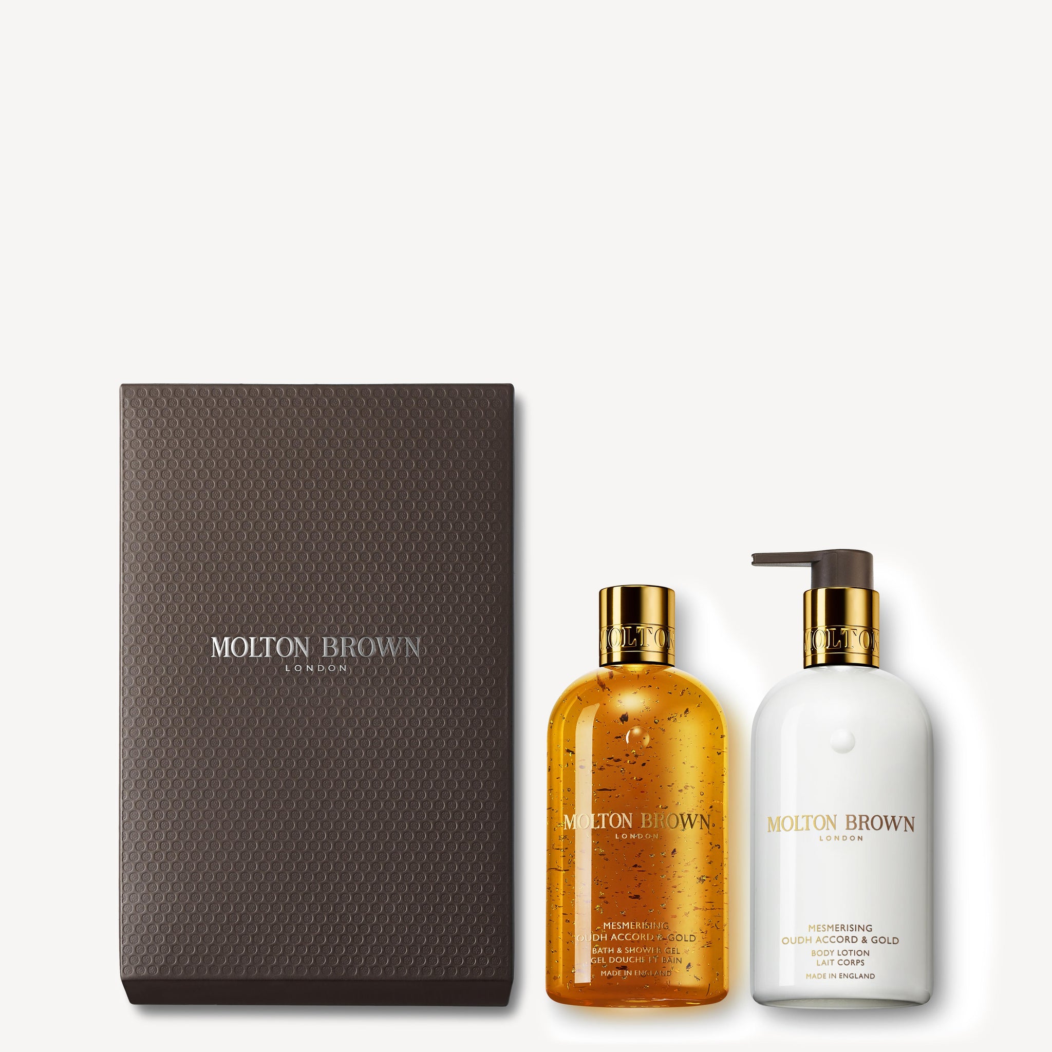 Introducing the Mesmerising Oudh Accord & Gold Shower Gel & Lotion Gift Set from MoltonBrown Cyprus, featuring a luxurious amber-colored Bath & Shower Gel with gold speckles and a white Oudh Accord Body Lotion with a pump. Each bottle is adorned with a gold cap and elegantly packaged in a textured dark brown box showcasing the MoltonBrown logo.