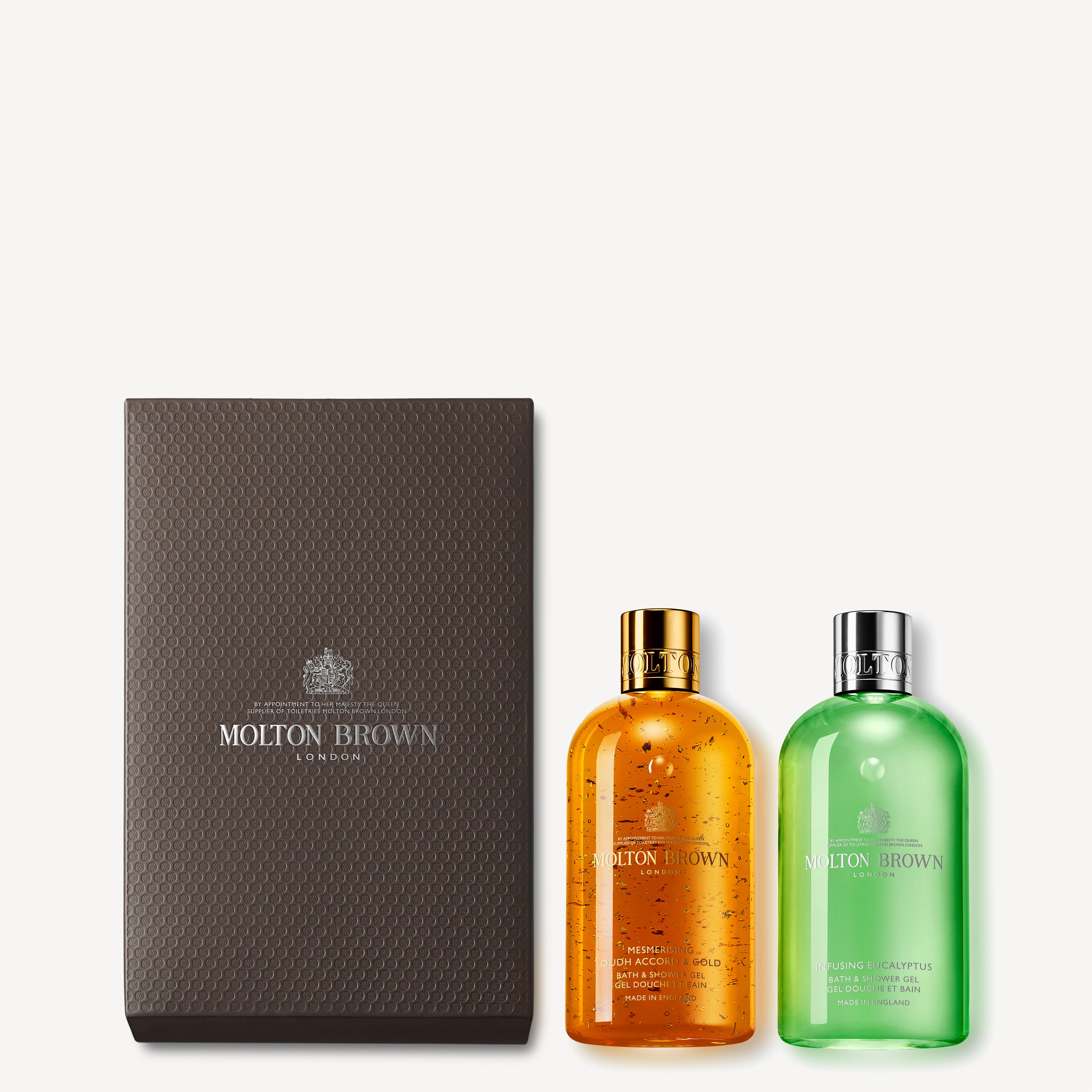 A MoltonBrown Cyprus gift box in a luxurious dark brown shade is elegantly displayed with the Mesmerising Oudh Accord & Gold & Eucalyptus Shower Gel Gift Set. The vibrant orange bottle, infused with eucalyptus and mint notes, boasts a gold cap, while the captivating green bottle gleams with 23.5-carat gold flakes beneath a sleek silver cap.