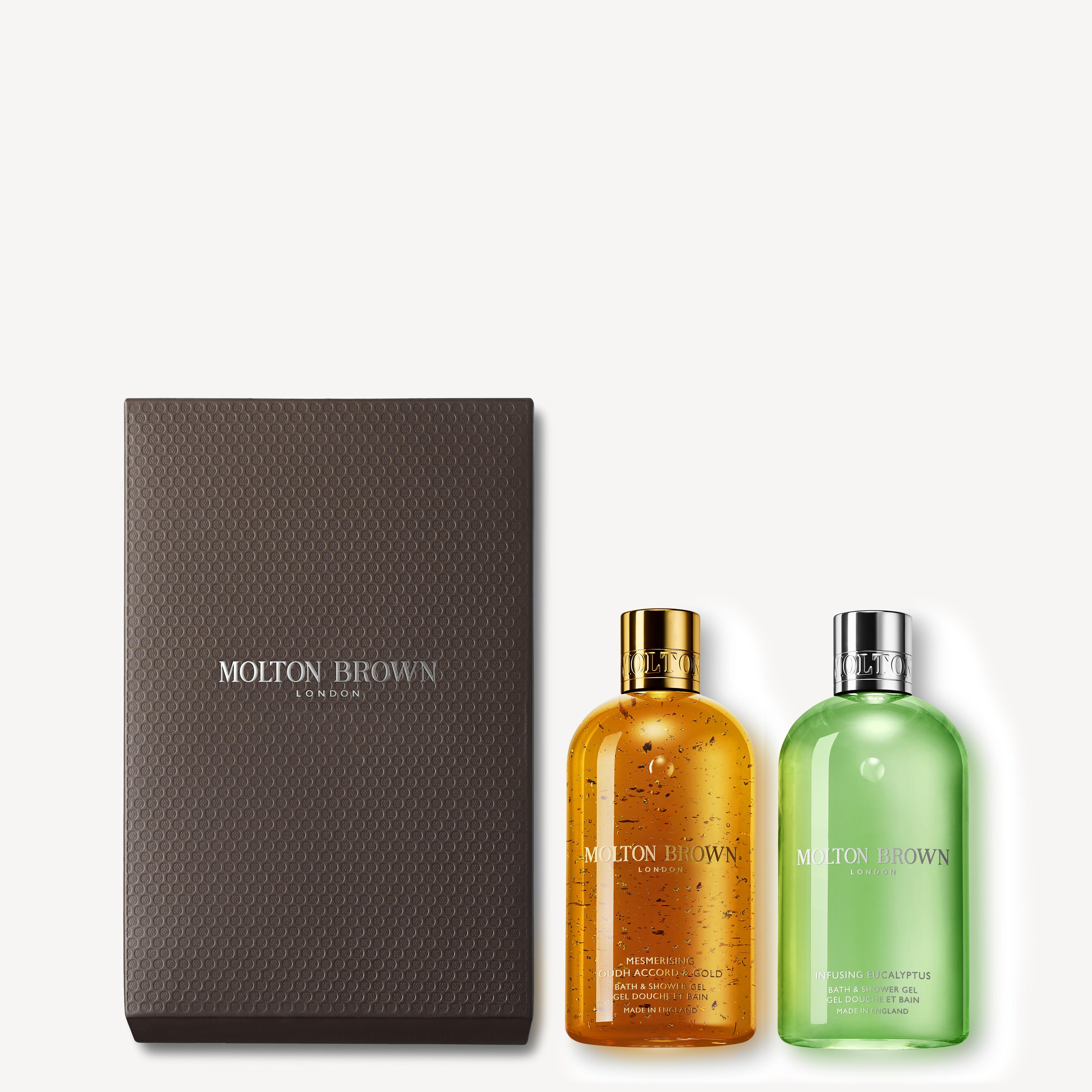 A MoltonBrown Cyprus gift box in a luxurious dark brown shade is elegantly displayed with the Mesmerising Oudh Accord & Gold & Eucalyptus Shower Gel Gift Set. The vibrant orange bottle, infused with eucalyptus and mint notes, boasts a gold cap, while the captivating green bottle gleams with 23.5-carat gold flakes beneath a sleek silver cap.