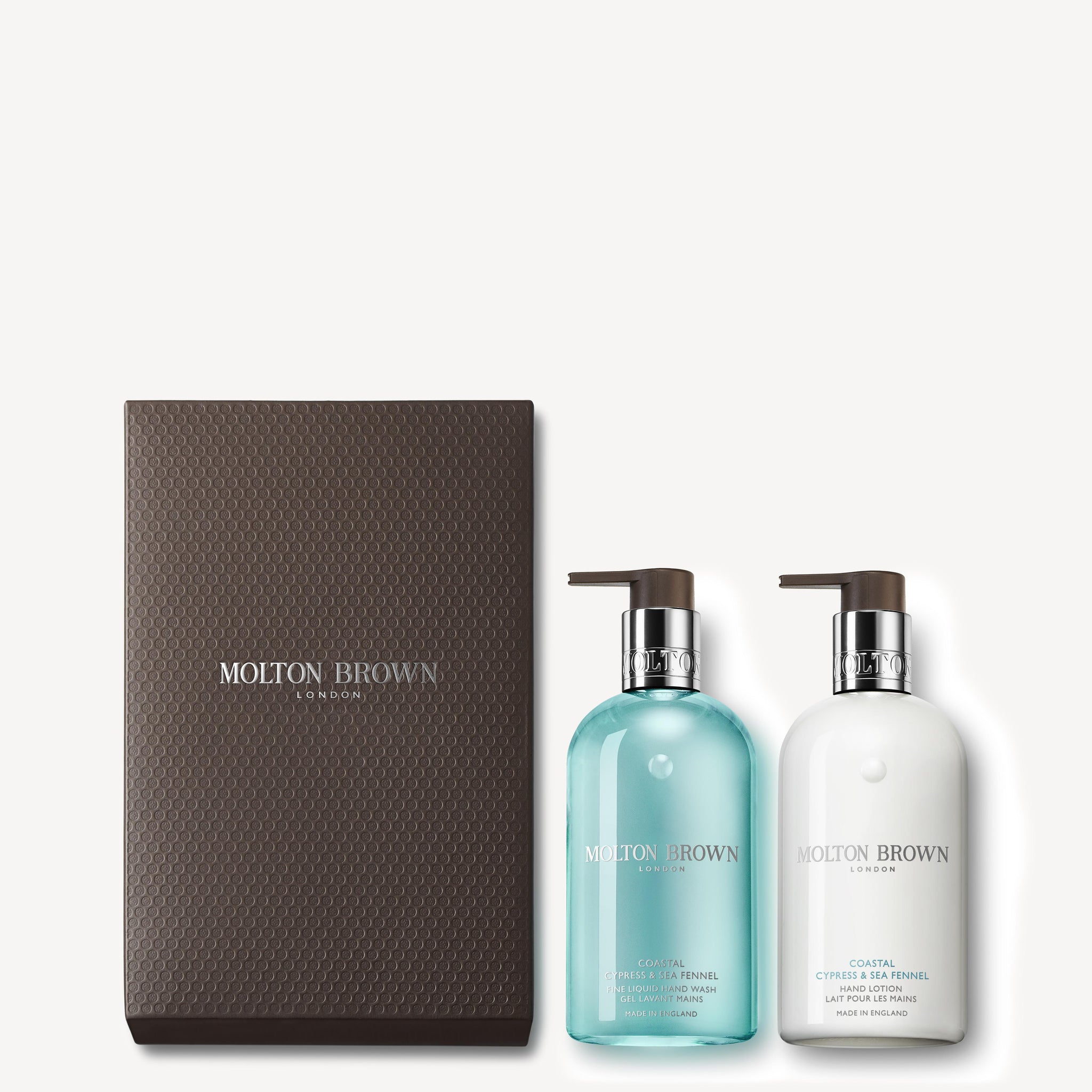 This Coastal Cypress & Sea Fennel Hand Wash & Lotion Set by MoltonBrown Cyprus is elegantly presented in a textured dark brown box. It includes two pump bottles: one blue and one white, offering a luxurious Fine Liquid Hand Wash and Hand Lotion for an indulgent sensory experience.