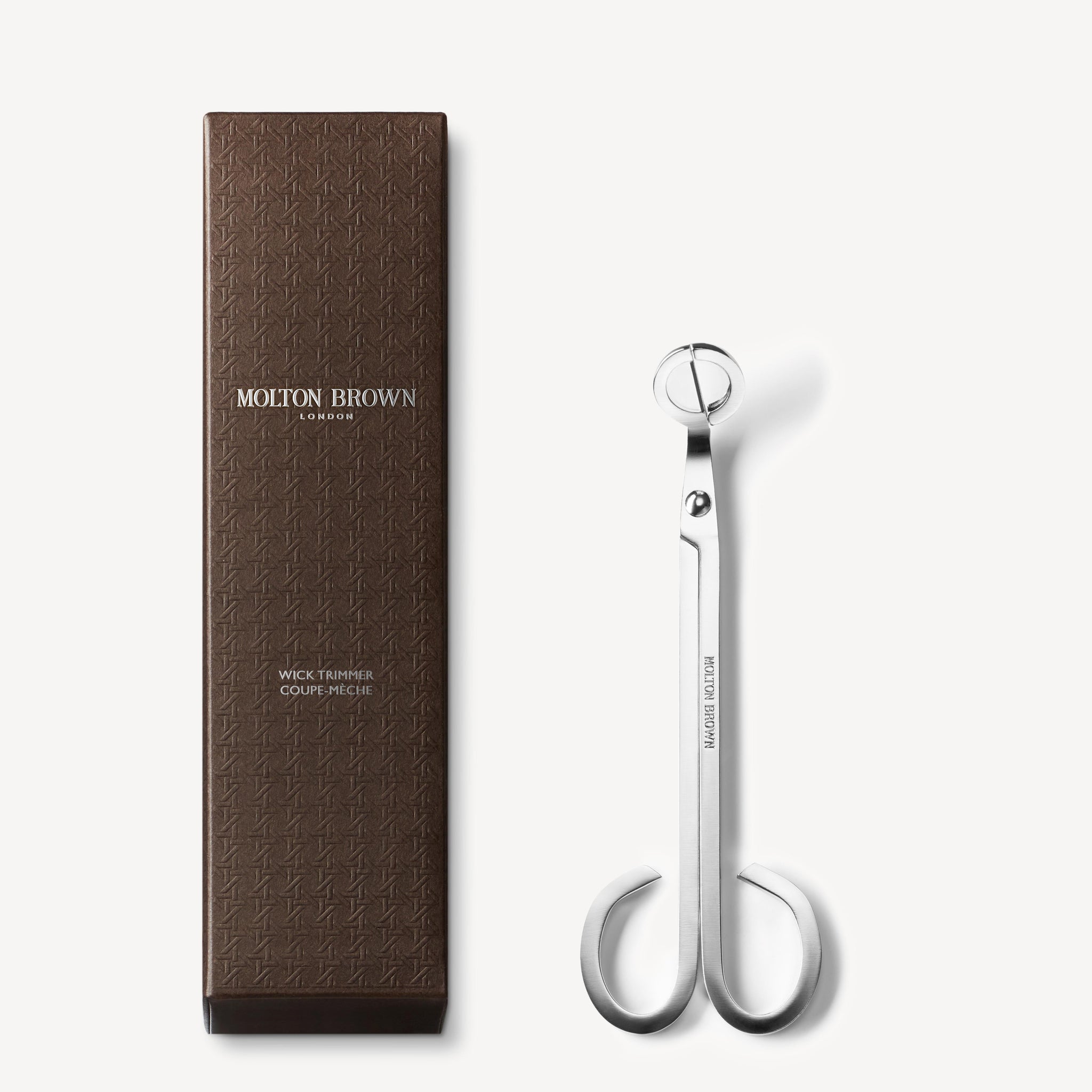 A pair of silver wick trimmers with a round handle, perfect for candle care, is elegantly displayed alongside a brown textured box labeled MoltonBrown Cyprus Wick Trimmer 72g, making it ideal for maintaining scented candles.